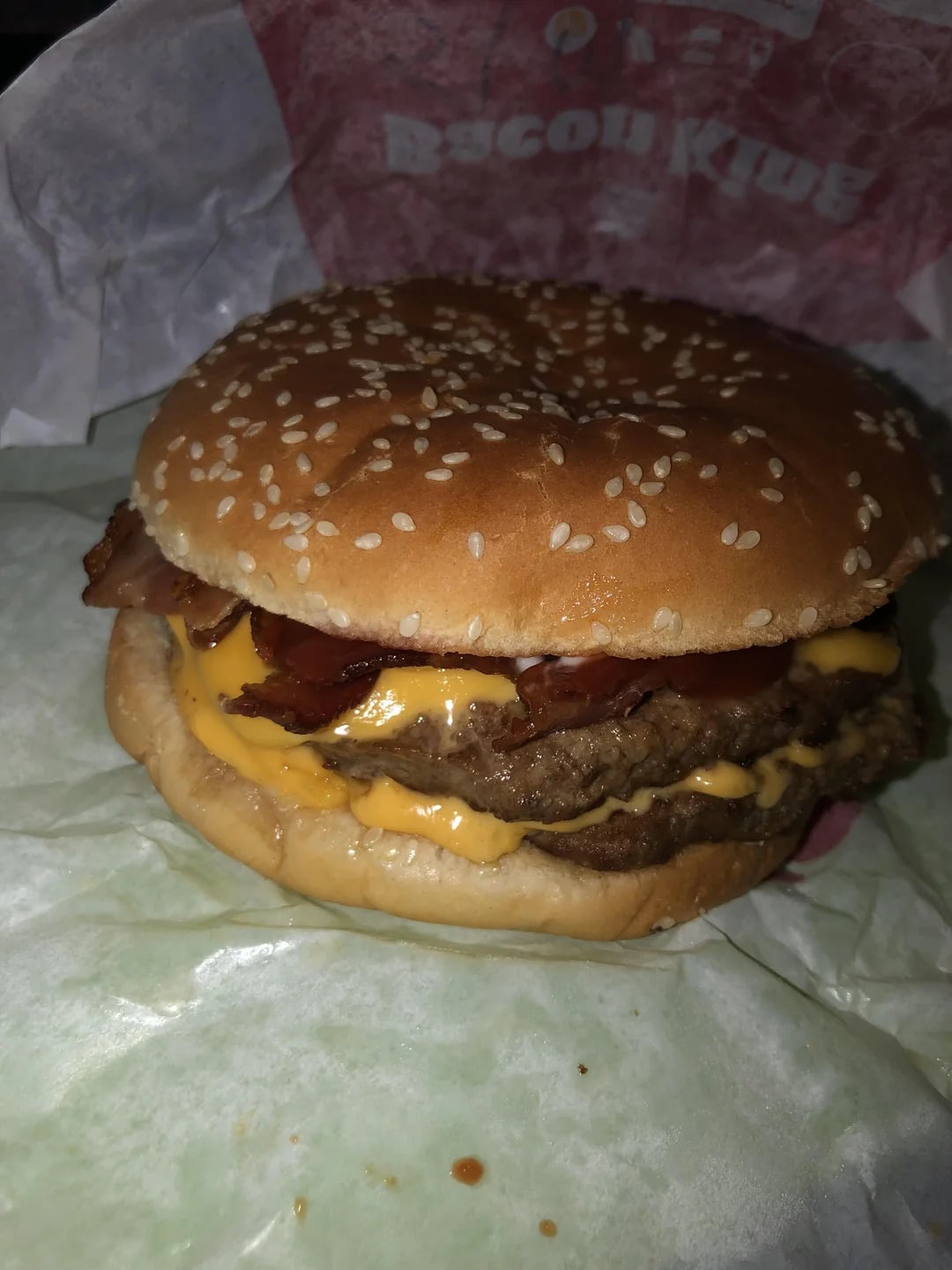 A close-up of a cheeseburger with a sesame seed bun, two beef patties, melted cheese, and crispy bacon, wrapped partially in branded fast food paper.