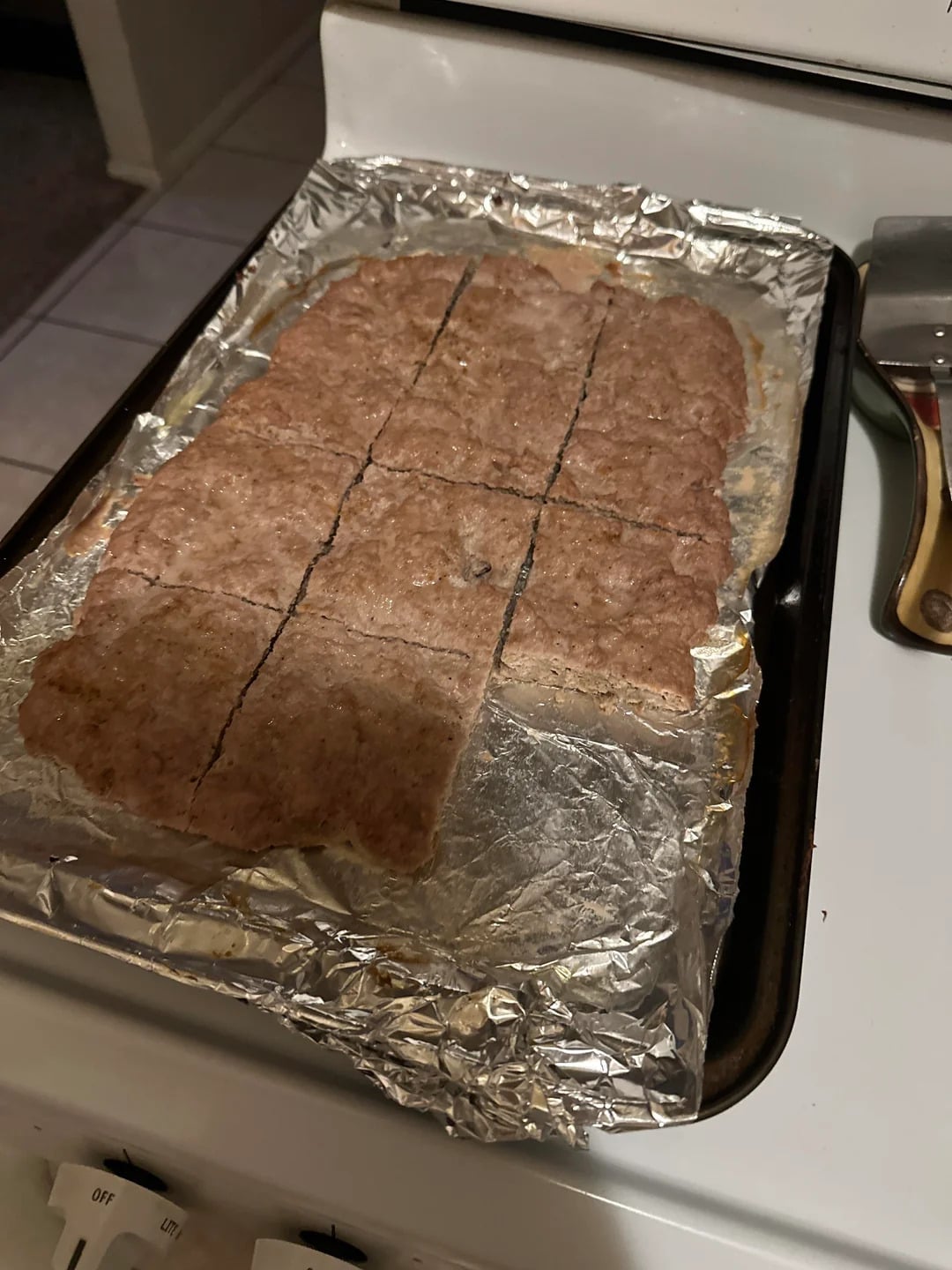 A baking sheet lined with foil holds a batch of baked, brown bars that have been cut into rectangles. A metal spatula rests nearby on the edge of the stovetop.
