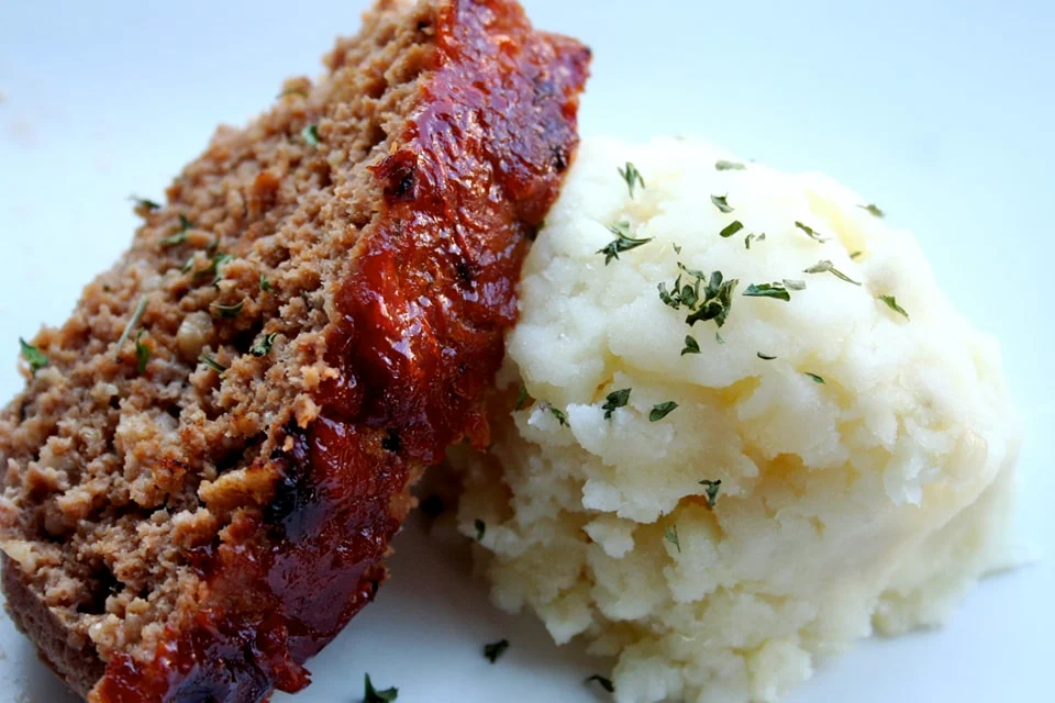 A slice of glazed meatloaf sits next to a serving of mashed potatoes, garnished with dried herbs, on a white plate.