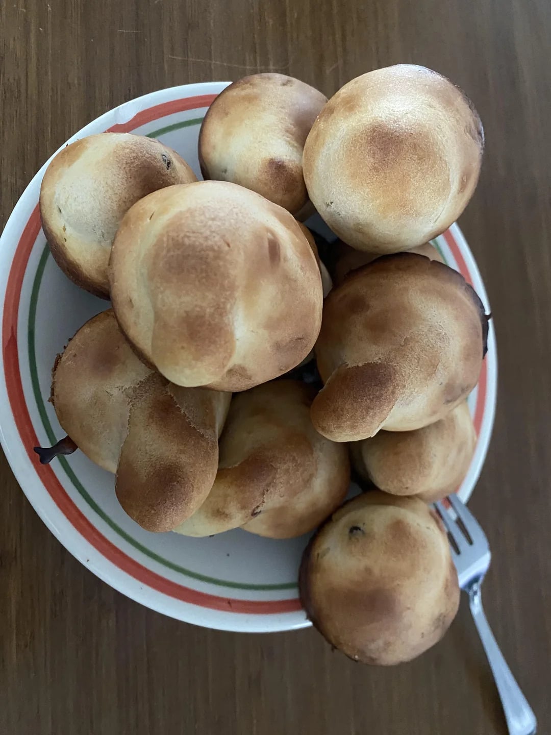 A plate with a pile of round, golden-brown baked rolls is shown on a wooden table. A fork rests on the side of the plate.