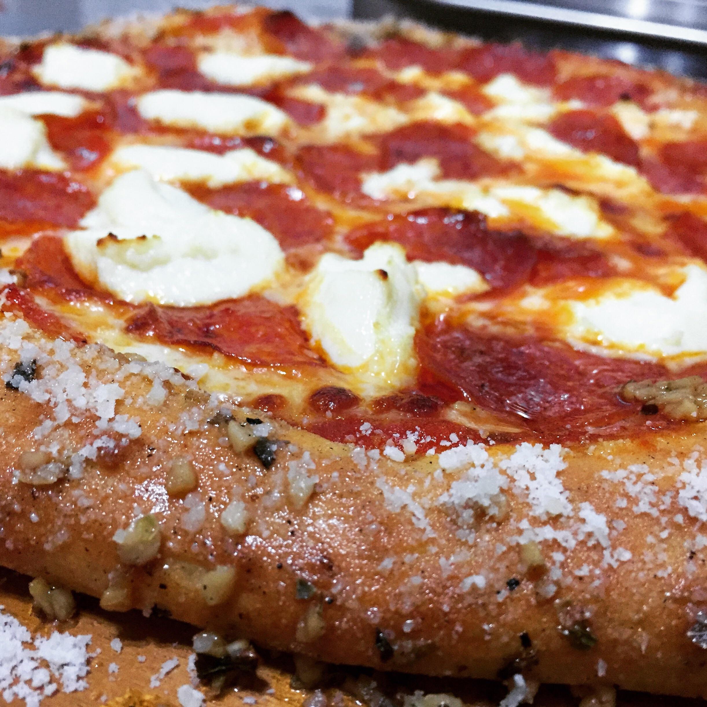 Close-up of a pepperoni pizza with dollops of ricotta cheese and a thick, seasoned crust topped with grated cheese and herbs.