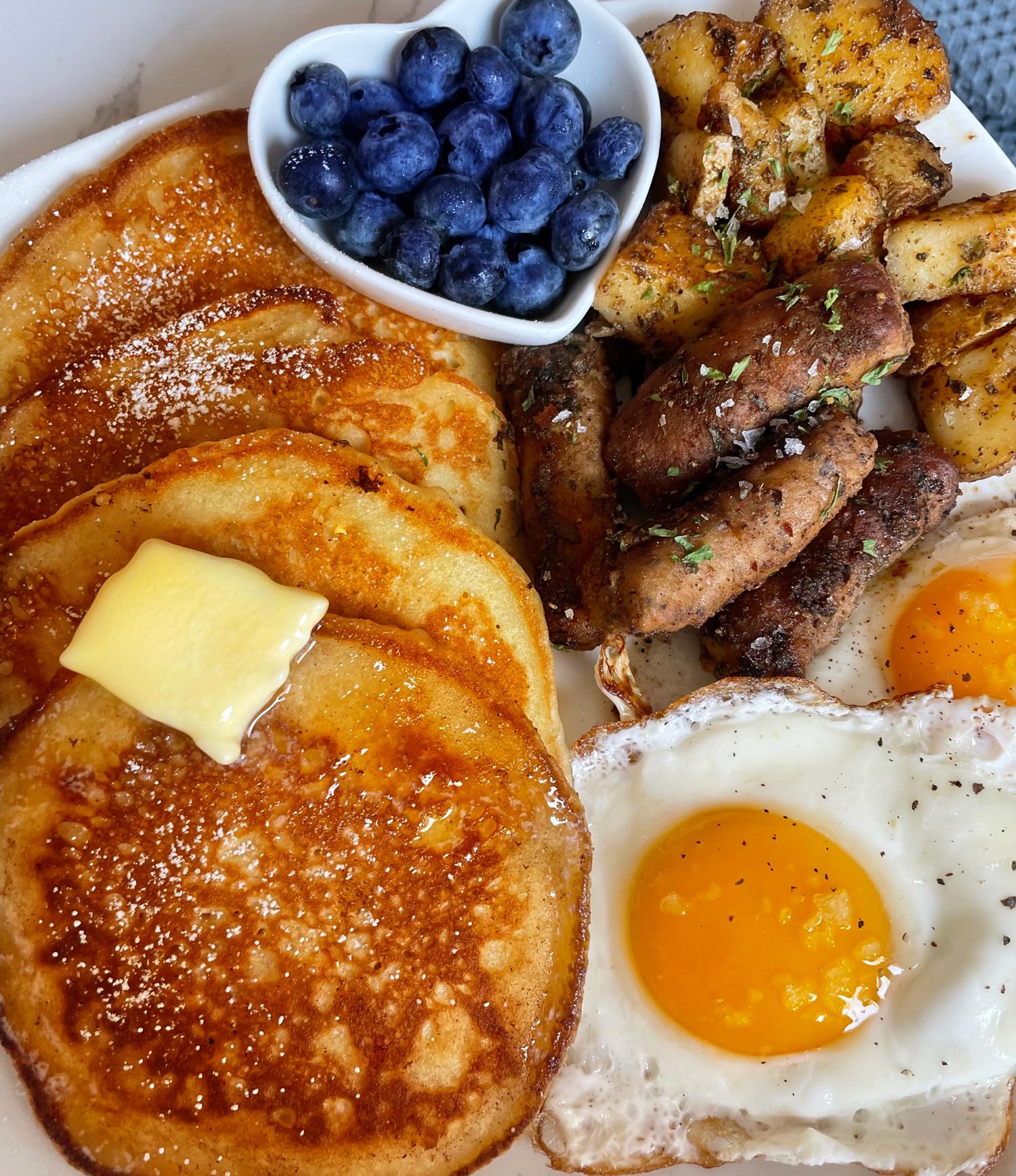 A breakfast plate featuring fluffy homemade pancakes topped with butter and syrup, sunny side up eggs, seasoned potatoes, sausage links, and a small dish of fresh blueberries.