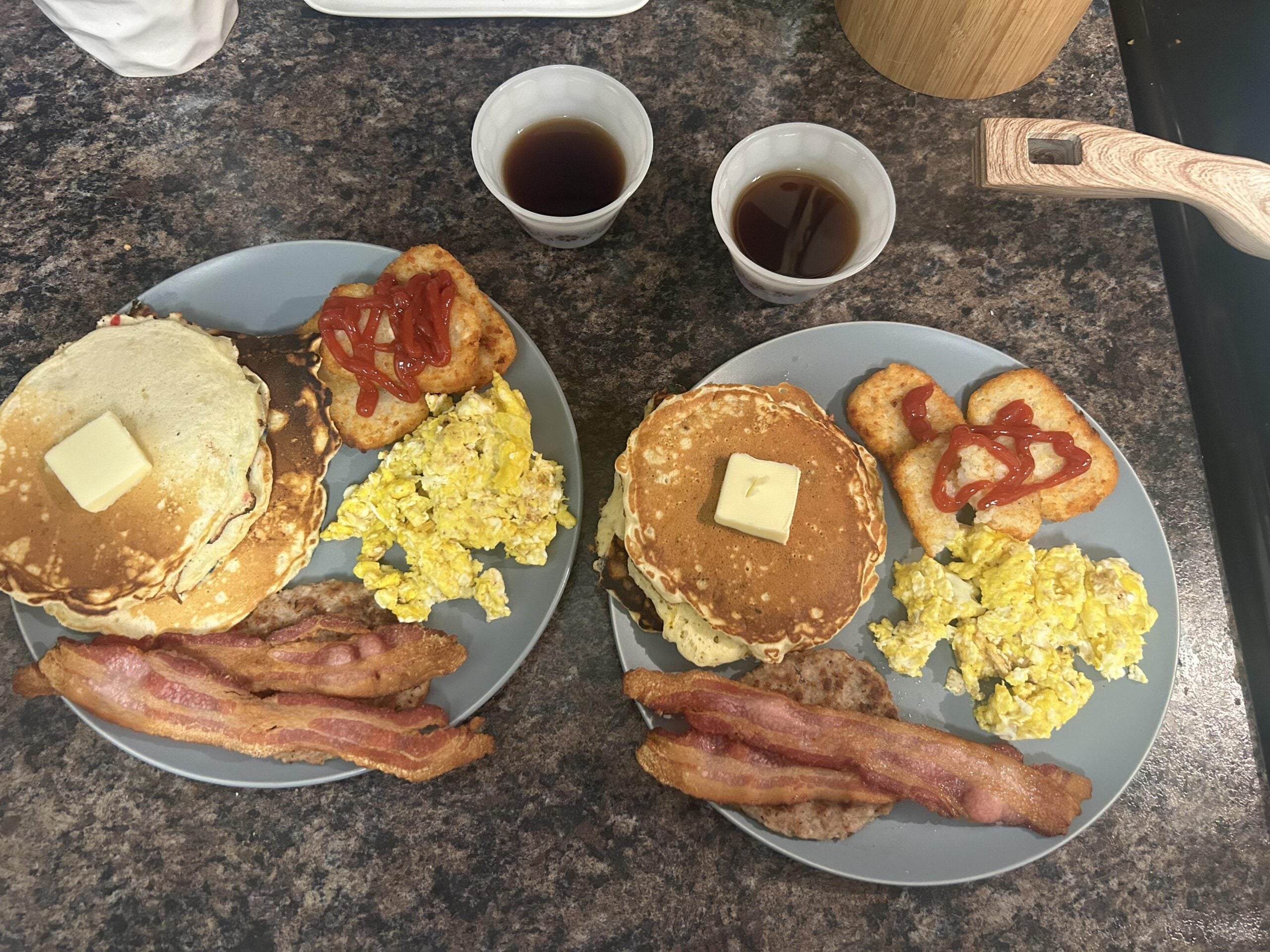 Two plates with pancakes topped with butter, scrambled eggs, bacon, sausage, hash browns with ketchup, and two cups of syrup on a dark countertop.