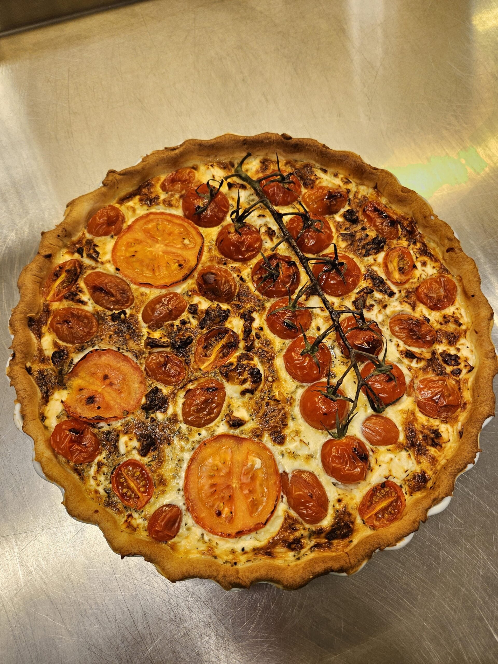 A baked quiche with a golden crust, topped with slices of tomato, halved cherry tomatoes, and a vine of roasted cherry tomatoes, on a metal countertop.