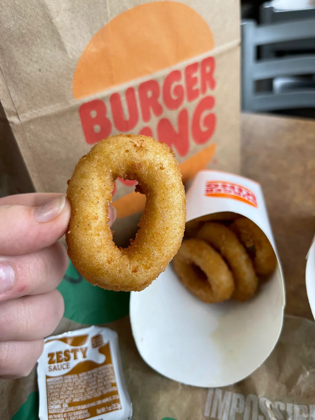 A hand holds up a golden-brown onion ring in front of a Burger King bag. More onion rings are in a white container nearby, along with a packet of Zesty sauce.