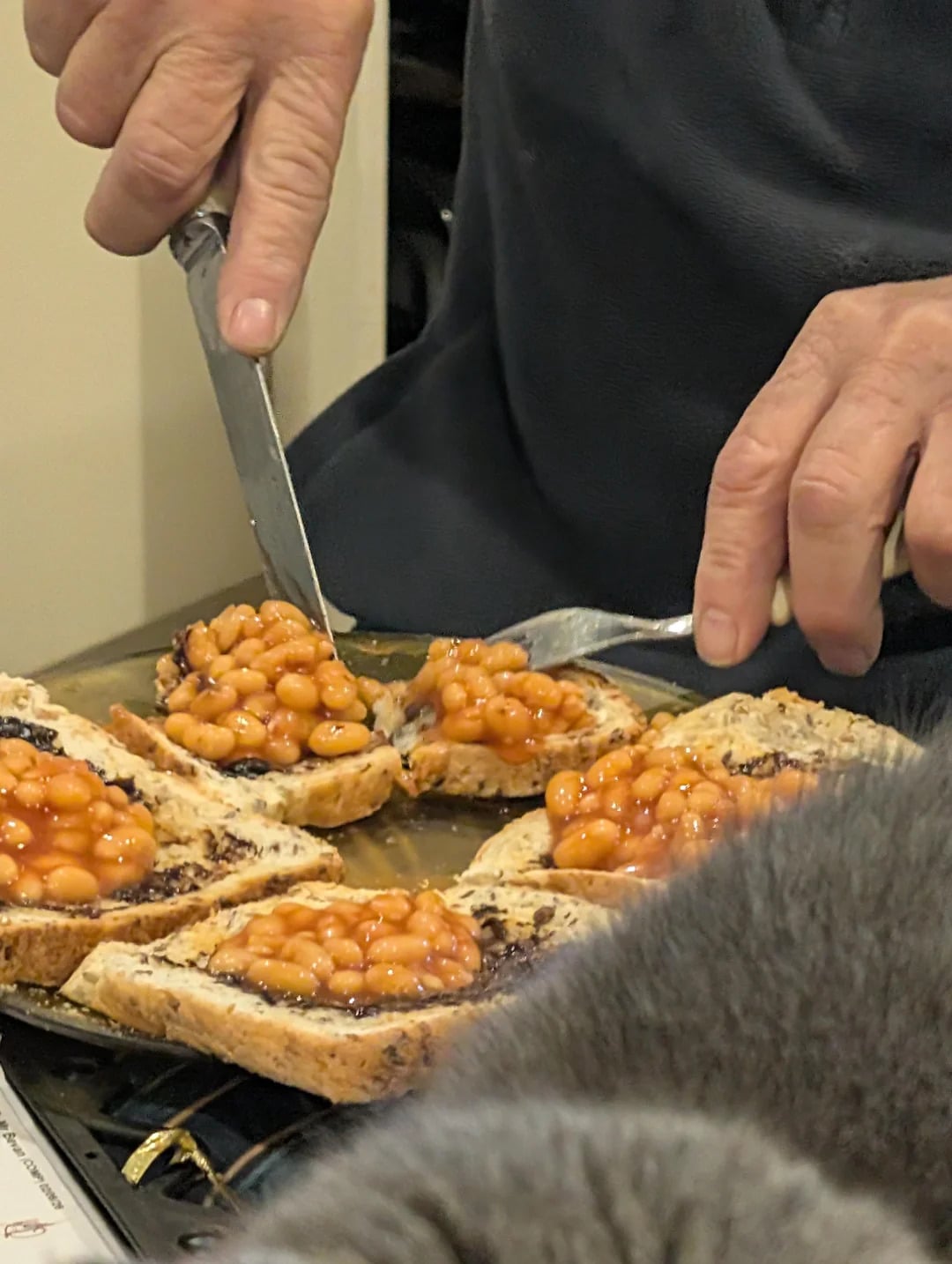 A person spreads baked beans on slices of bread using a knife and fork, preparing multiple servings on a countertop.