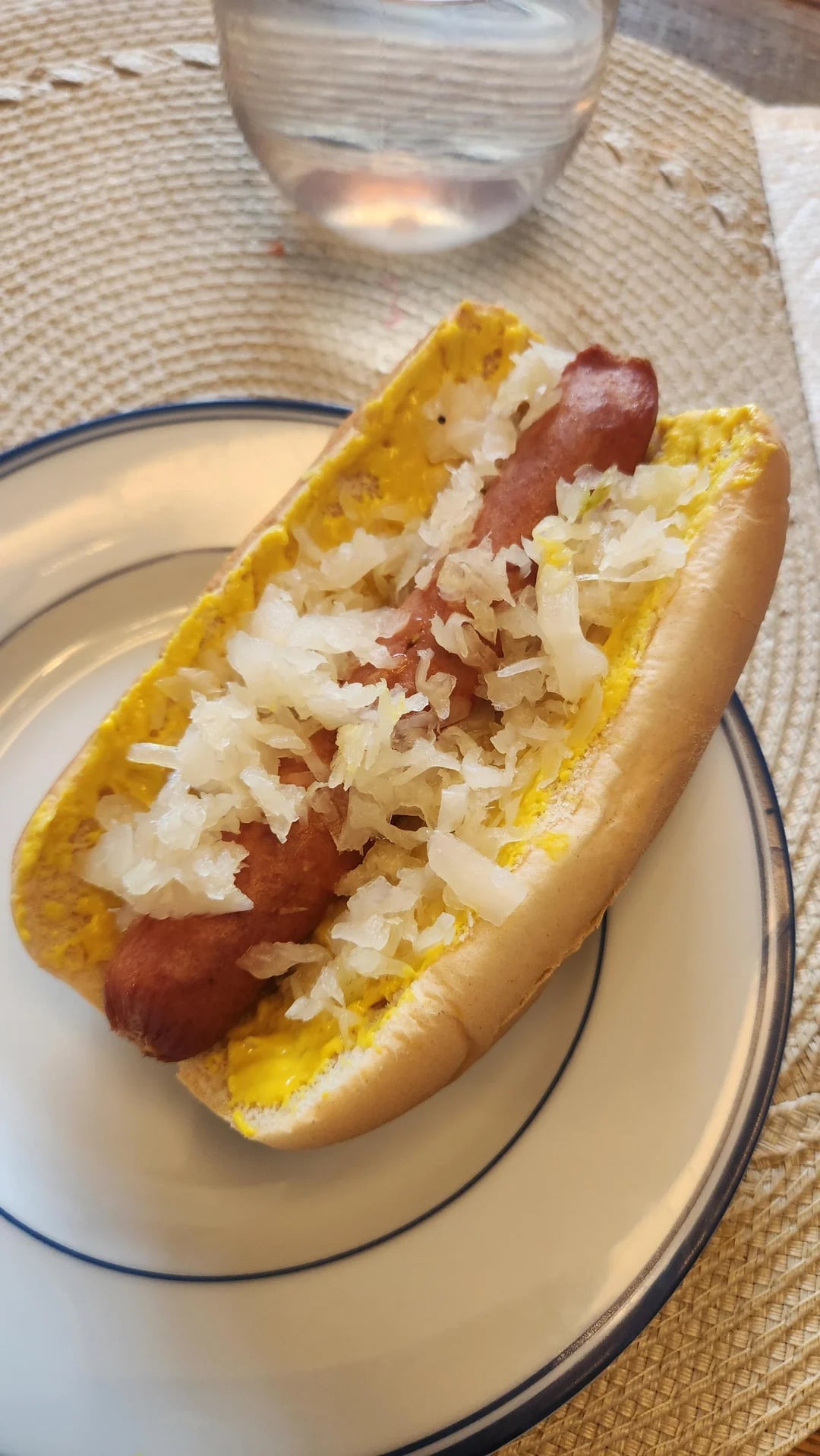 A hot dog in a bun topped with mustard and sauerkraut sits on a white plate with blue trim, placed on a woven placemat. A glass of water is visible in the background.