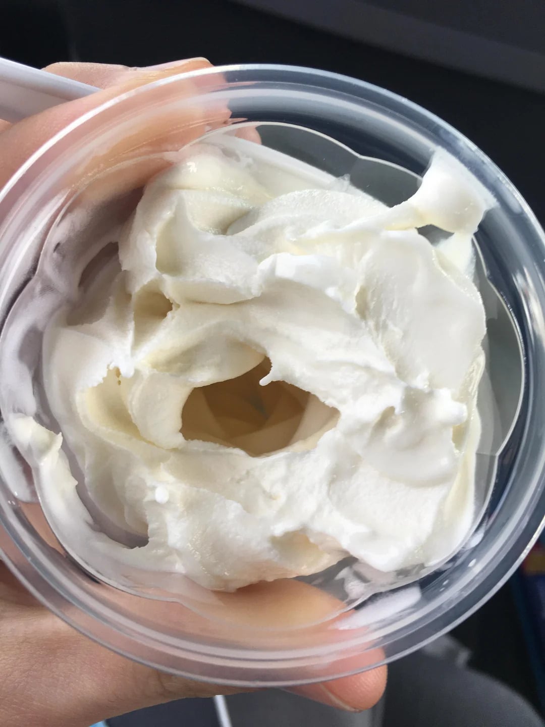 A hand holding a clear plastic cup filled with thick, white whipped cream or yogurt, with a visible hollow center.
