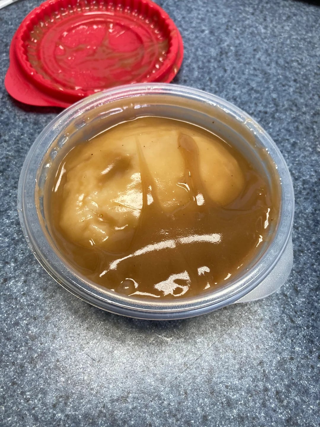 A plastic container filled with mashed potatoes and brown gravy, reminiscent of fast-food grocery store products, sits on a gray countertop. A red lid with some gravy residue is nearby.