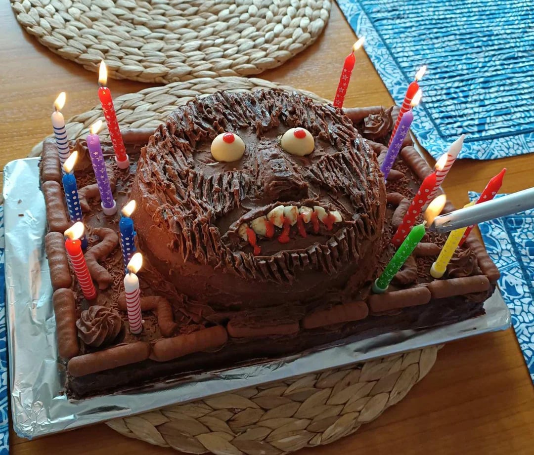 A chocolate birthday cake decorated to look like a monster face, with candy eyes and teeth, surrounded by lit colorful candles on a decorated table.