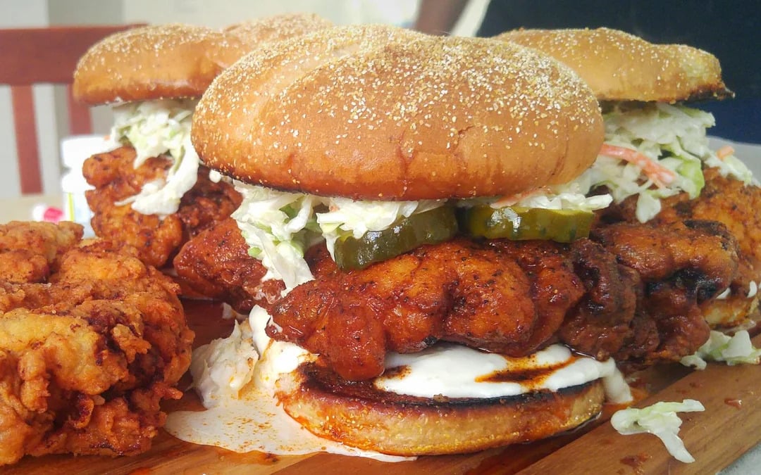Two fried chicken sandwiches with slaw, pickles, and sauce on buns. A piece of fried chicken sits on the side. The sandwiches are on a wooden board, and the background shows a chair.
