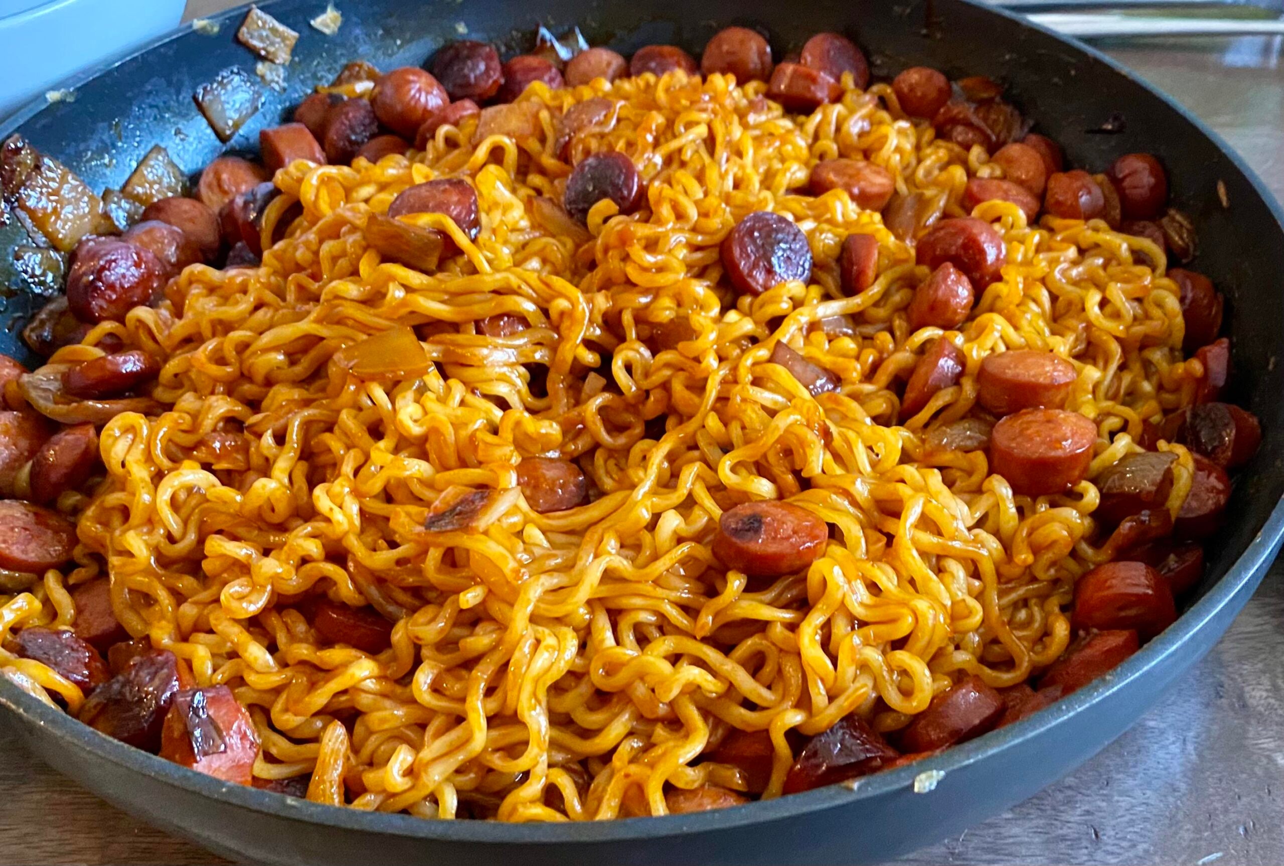 A pan filled with cooked ramen noodles mixed with sliced sausages and chunks of browned onions in a savory sauce.