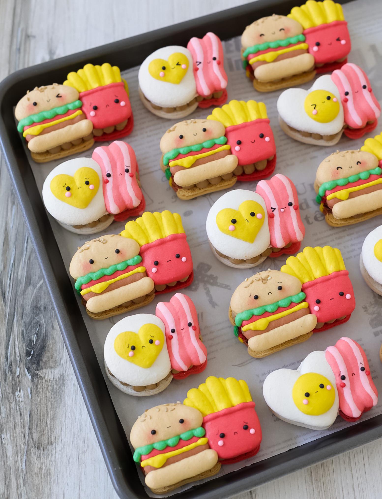 A tray of colorful macarons shaped like burgers, fries, eggs, and bacon, each with cute smiling faces, displayed on parchment paper.