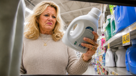 A woman with blonde hair in a beige sweater looks closely at a white detergent bottle in a store aisle, appearing concerned or confused by the label. Shelves of name-brand products and other cleaning items are visible around her.