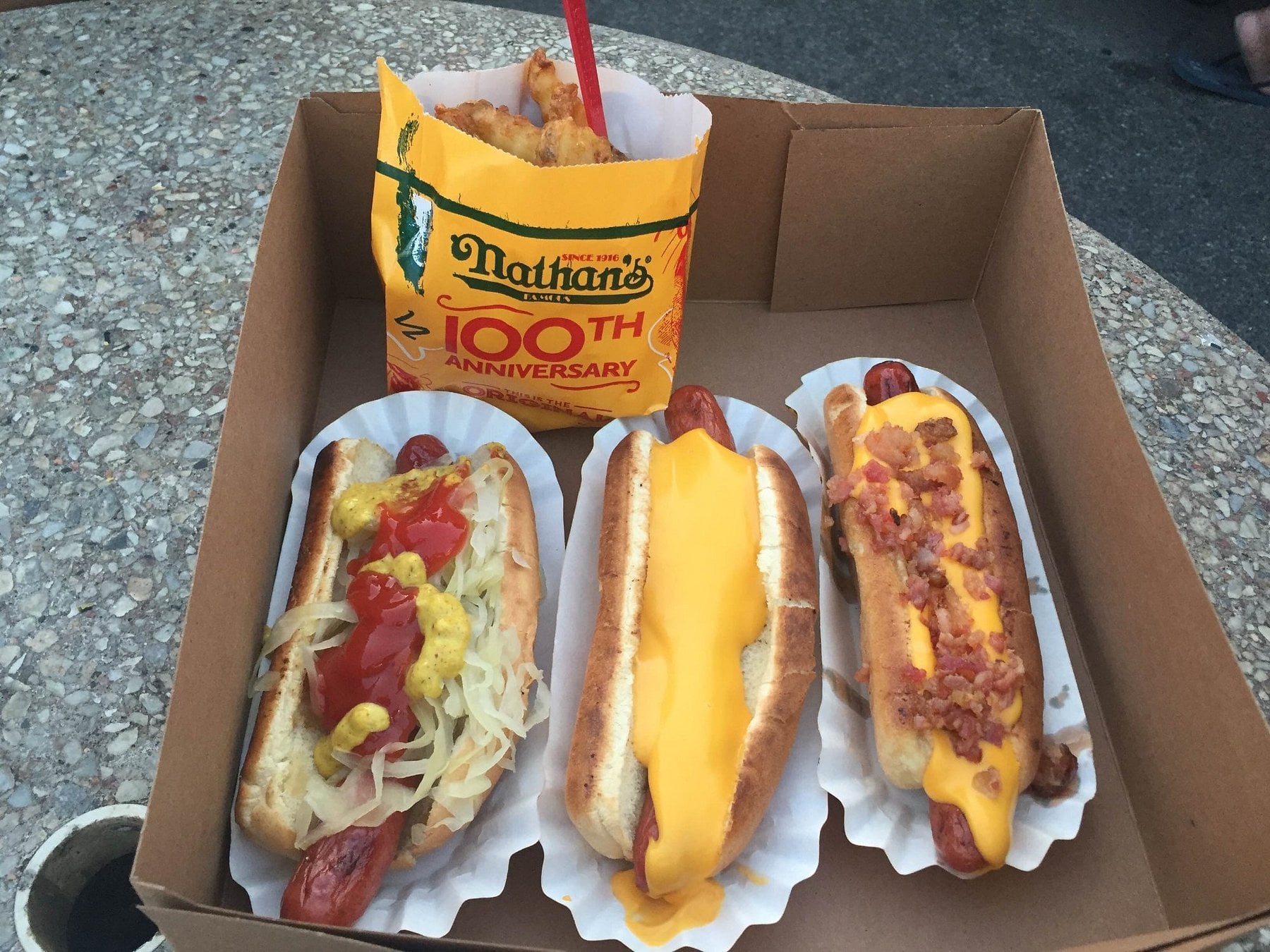 Three hot dogs in paper trays, each with different toppings: ketchup and sauerkraut, melted cheese, and cheese with bacon. A container of Nathan’s French fries is behind them, all on a cardboard tray on a stone table.