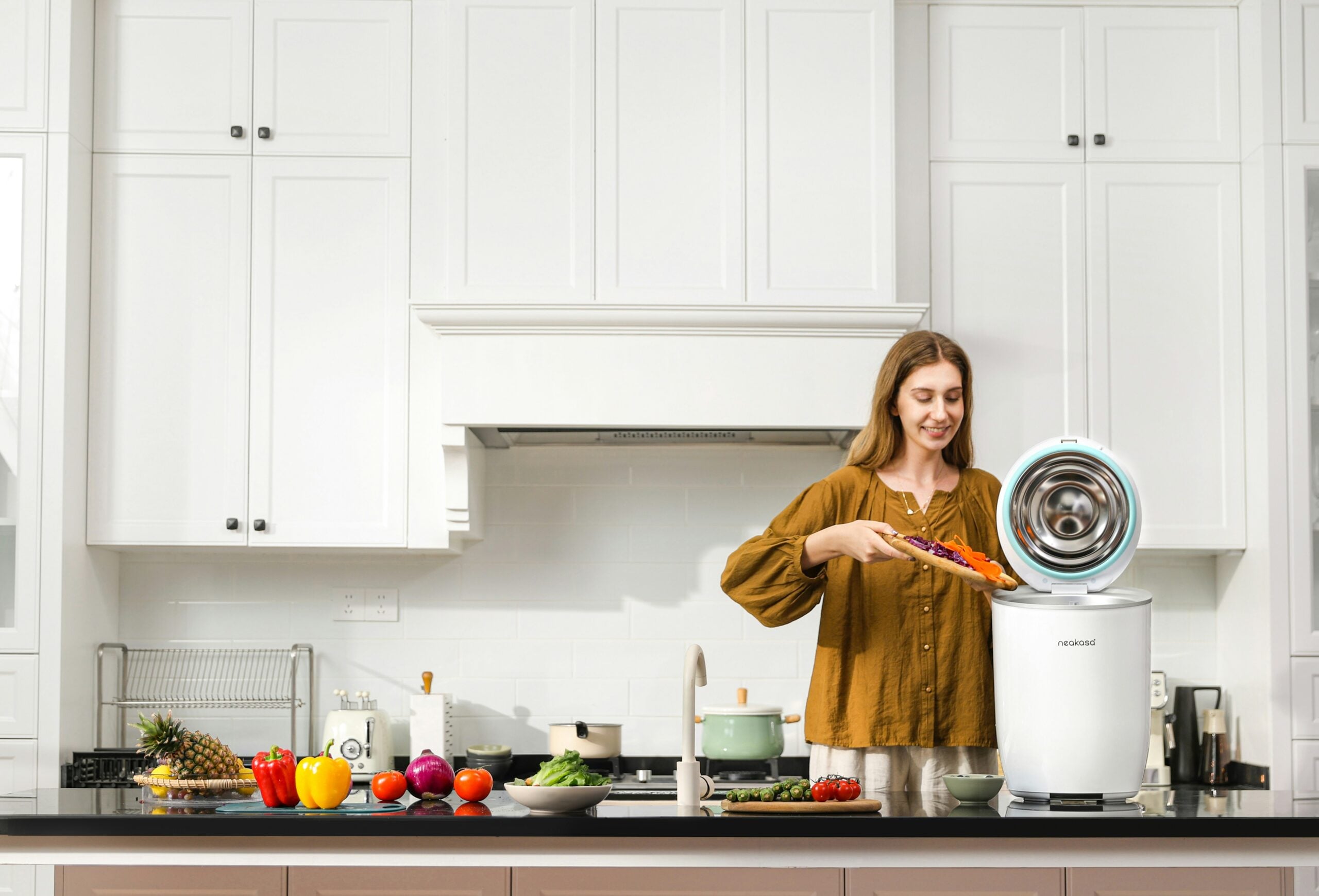 A woman in a kitchen places vegetable scraps into a white food waste compost bin on the counter. Fresh produce and kitchen utensils are arranged neatly around her.