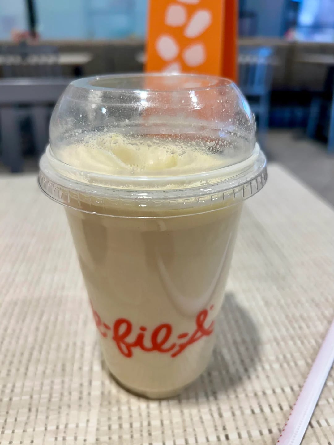 A vanilla milkshake in a clear plastic cup with a dome lid, featuring part of the Chick-fil-A logo, sits on a table with a blurred background, evoking the classic charm of fast-food grocery store products.