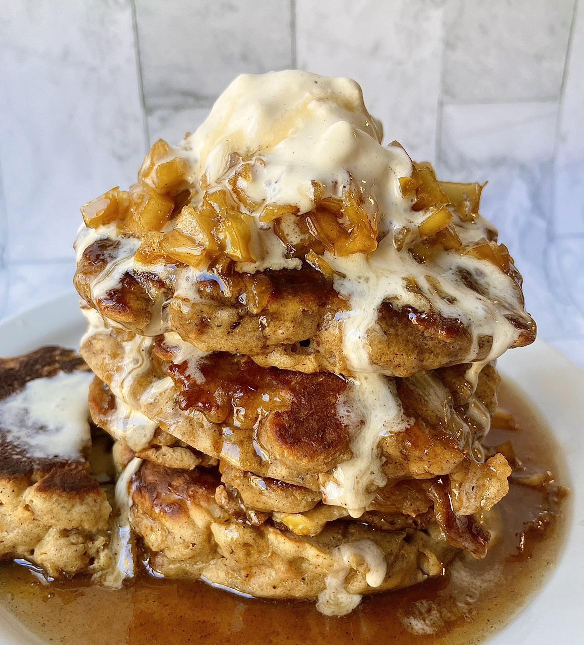 A tall stack of homemade pancakes topped with chopped caramelized apples, syrup, and a scoop of melting vanilla ice cream sits on a white plate against a light background.