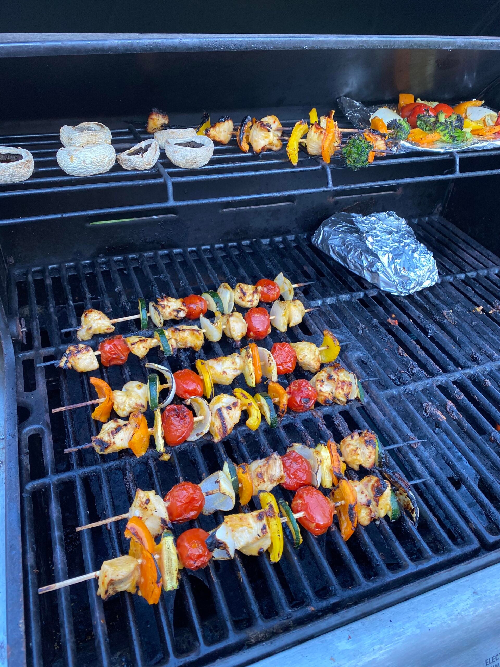 Colorful skewers with chicken, tomatoes, and peppers grill on the bottom rack—just what you need for the best BBQ cookouts this summer. The top rack holds mushroom caps, bread slices, peppers, and broccoli while a foil-wrapped item sits at the back. Everything is cooking to perfection on a barbecue grill.