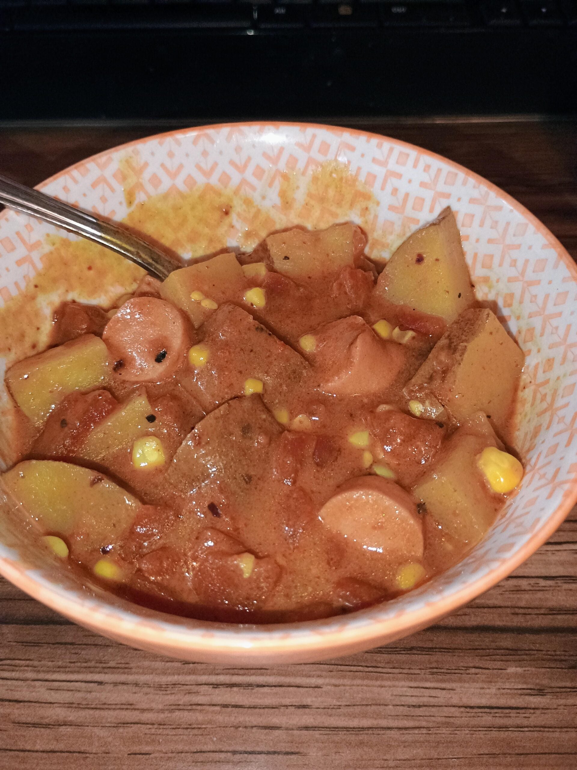 A bowl of stew containing sliced sausages, potatoes, corn, and tomato-based broth—thankfully spared from any hilarious cooking disasters—with a spoon inside the bowl. The bowl features an orange geometric pattern.