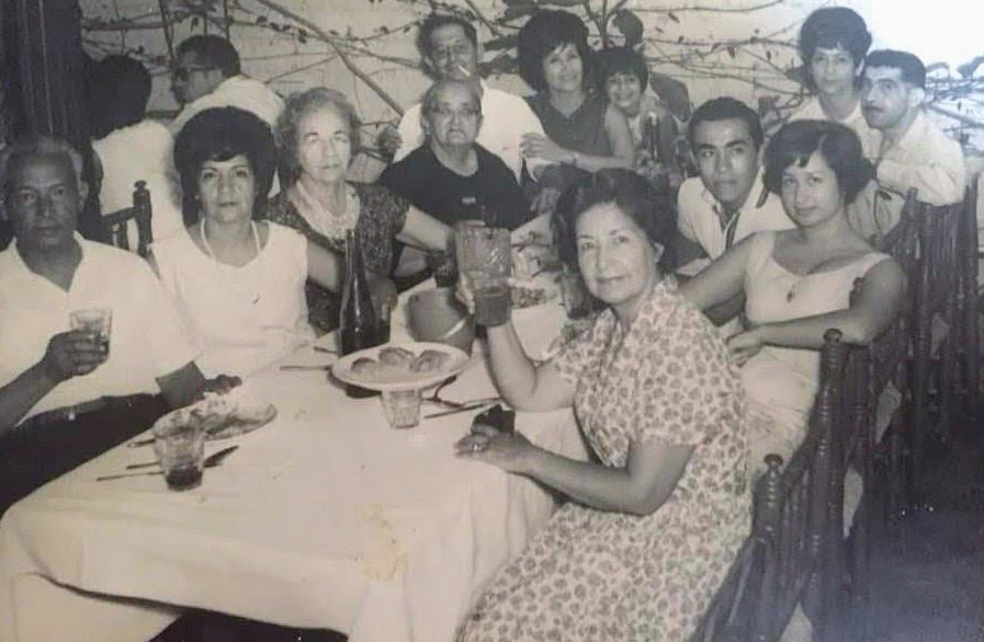 A group of men and women sit around a dining table, smiling and raising glasses. The photo is in black and white, suggesting it is from a past decade. There is food on the table, evoking the warmth of nostalgic dinners, and the setting appears festive and joyful.