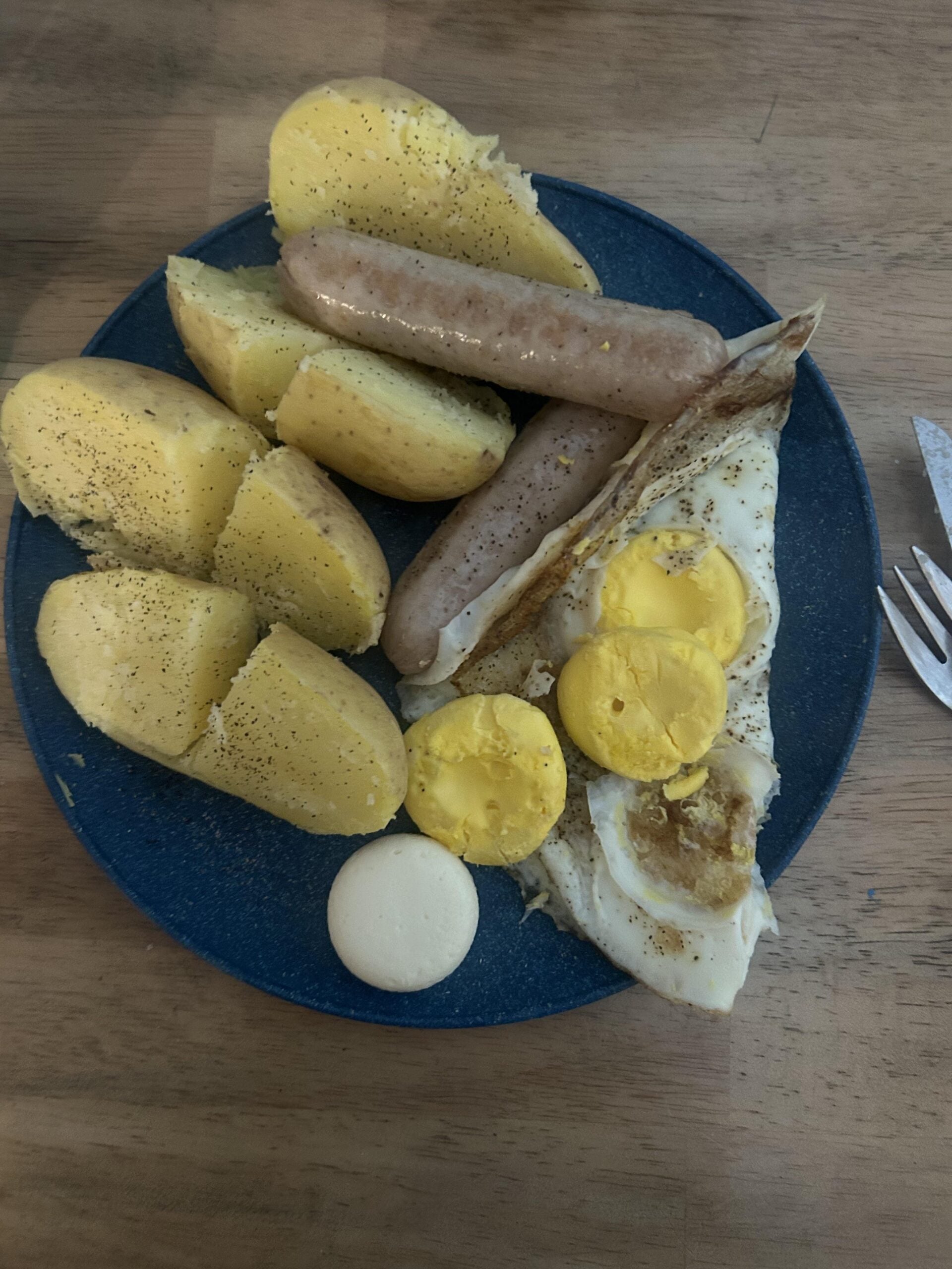 A blue plate with boiled potatoes, sausages, a fried egg with broken yolk, and a dollop of mayonnaise, all on a wooden table. A fork is partially visible to the right.