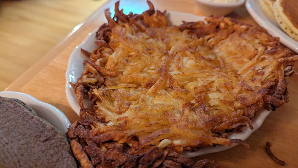 A close-up of a crispy, golden-brown hash brown served on a white plate, placed on a wooden table with other food items partially visible around it.