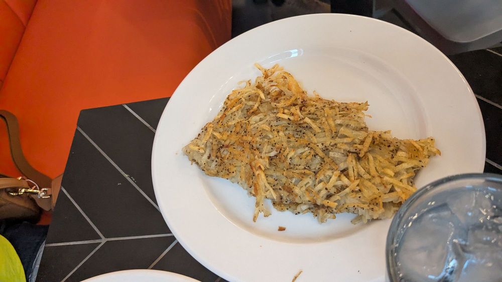 A white plate of crispy, seasoned hash browns sits on a dark, patterned table next to a glass of ice water. An orange seat and part of a brown bag are visible in the background.
