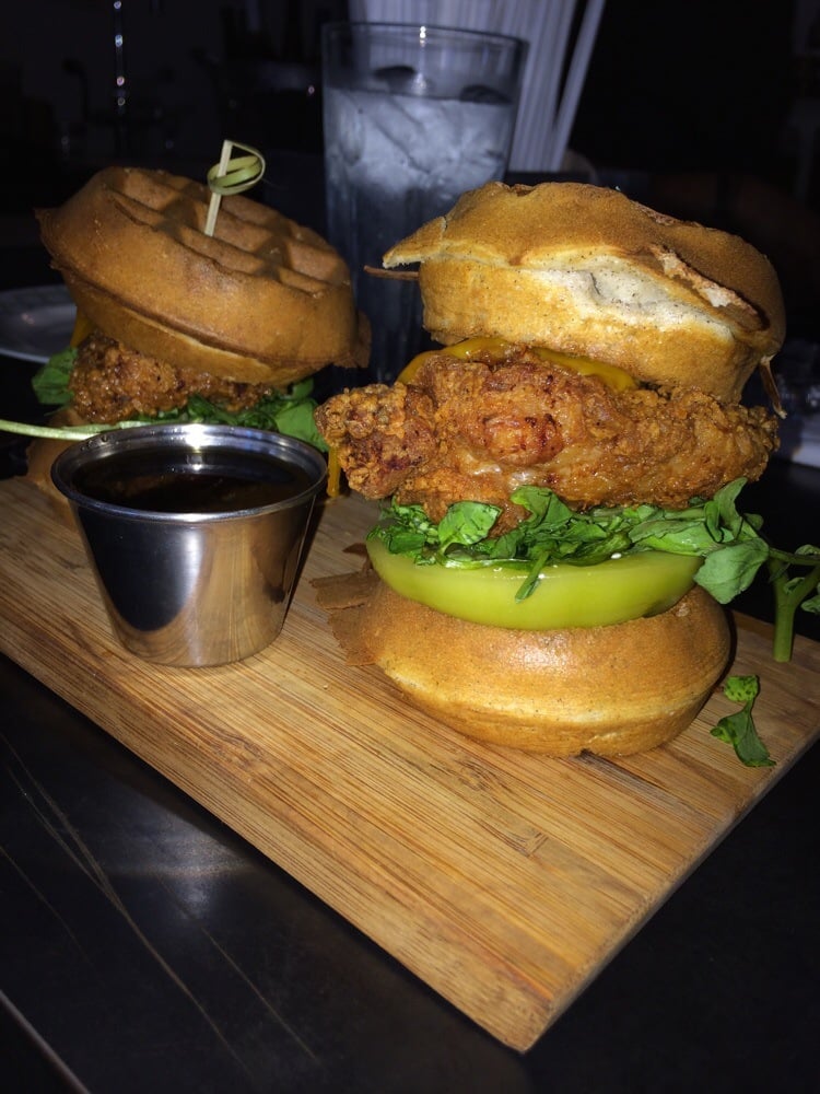 Two waffle sandwiches with fried chicken, lettuce, and tomato are served on a wooden board with a small cup of syrup. A glass of ice water is in the background.
