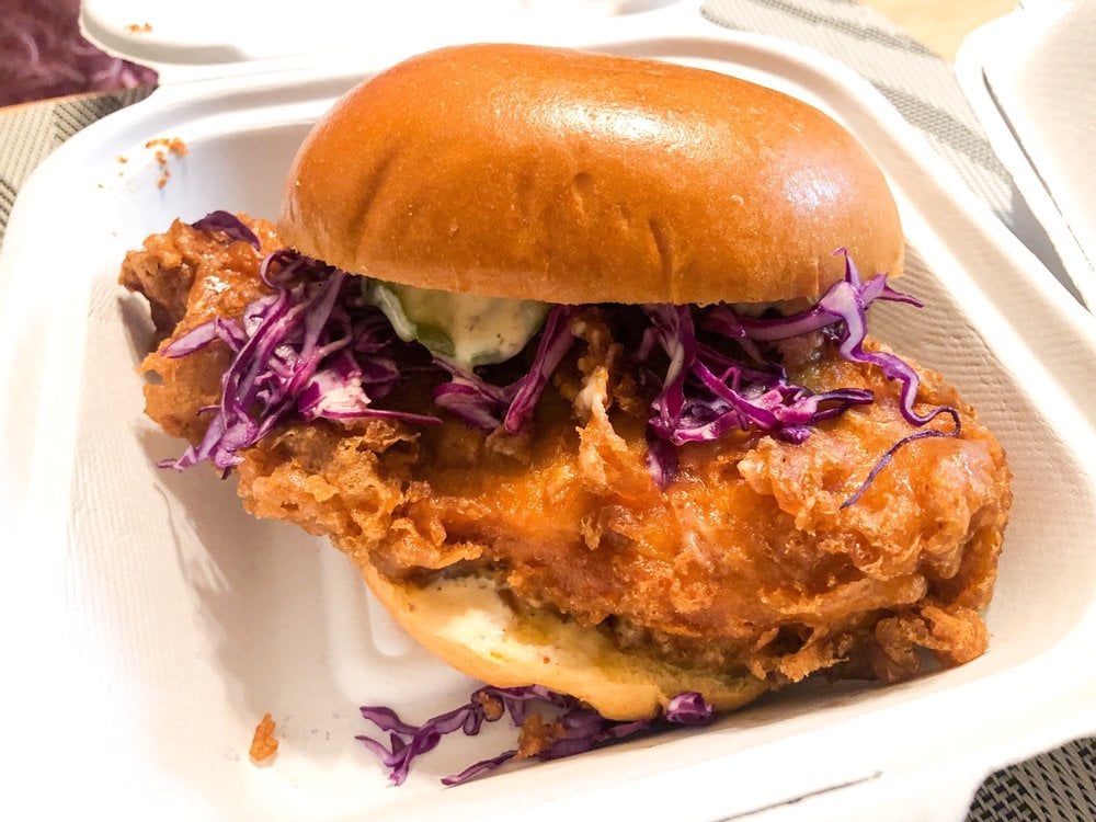 A crispy fried chicken sandwich with purple cabbage slaw and sauce, served on a soft bun in a white takeout container.