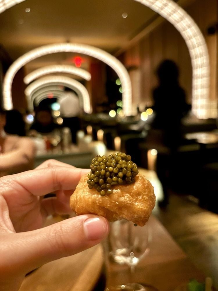 A hand holds up a small pastry topped with black caviar in an elegant, warmly lit restaurant with arched ceiling lights and blurred diners in the background.