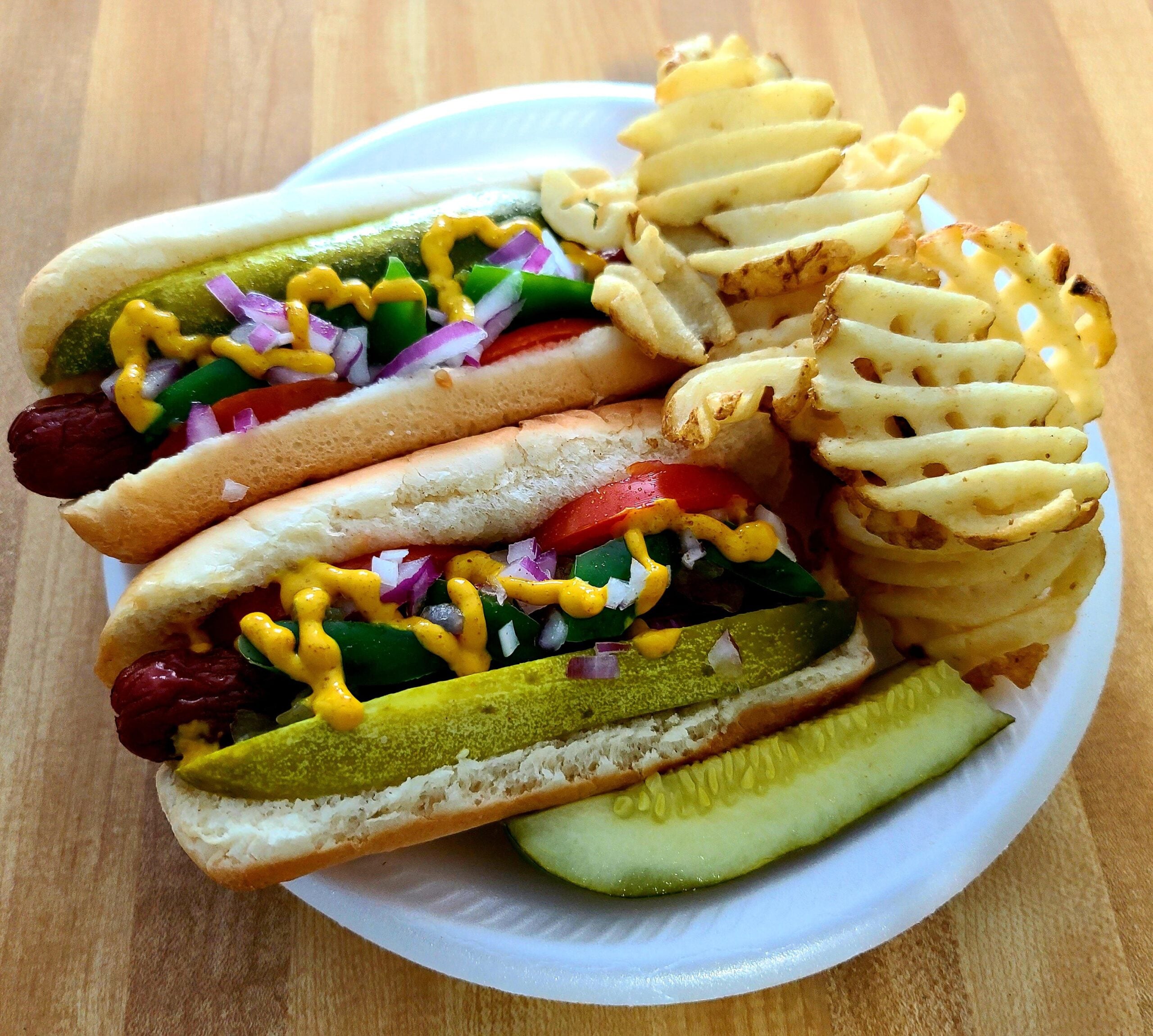 Two hot dogs with pickles, onions, tomato, and mustard, served on a white plate with crinkle-cut waffle fries and a pickle spear on a wooden table.