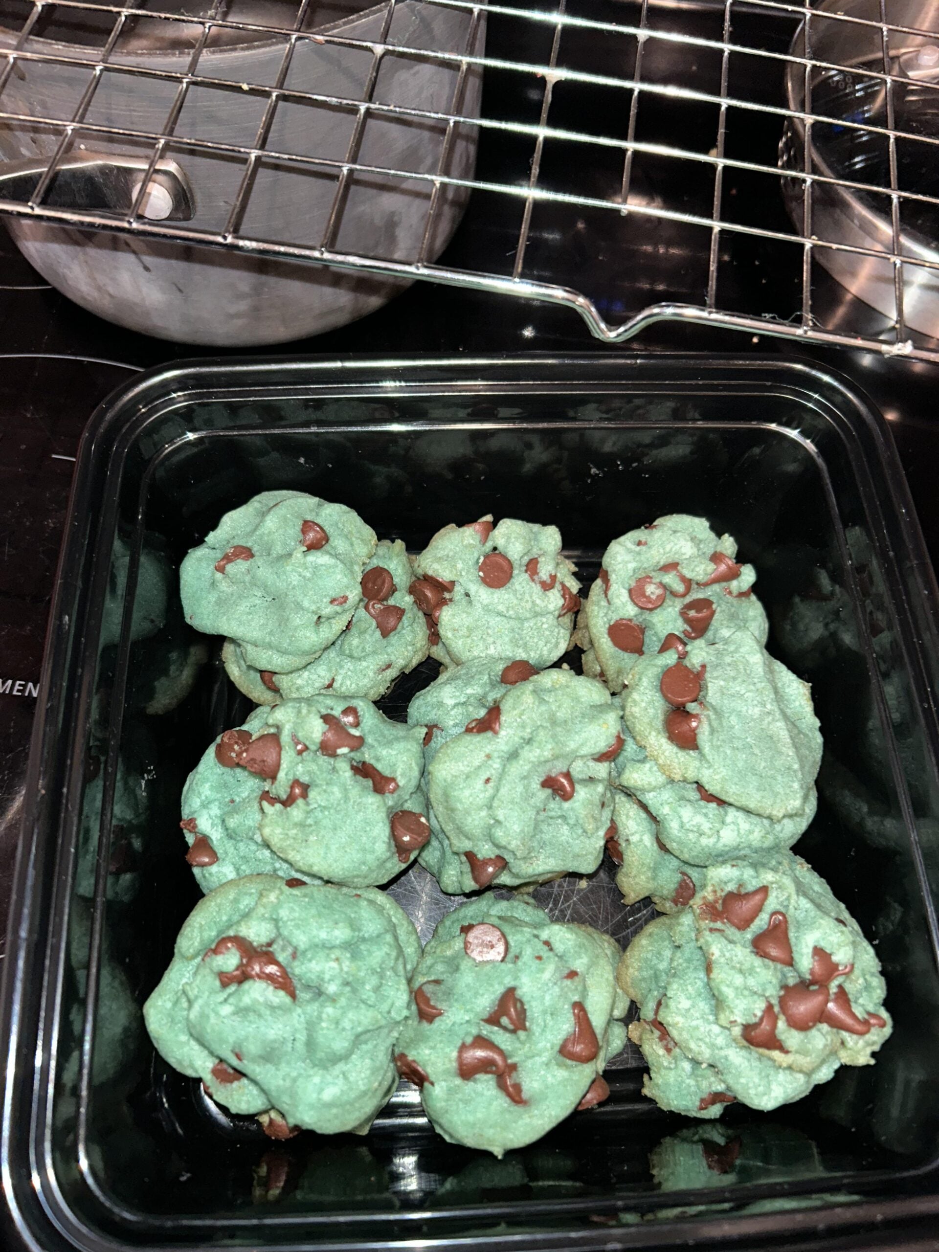 A container of green chocolate chip cookies sits on a stove, with a metal pot and an empty cooling rack visible in the background.