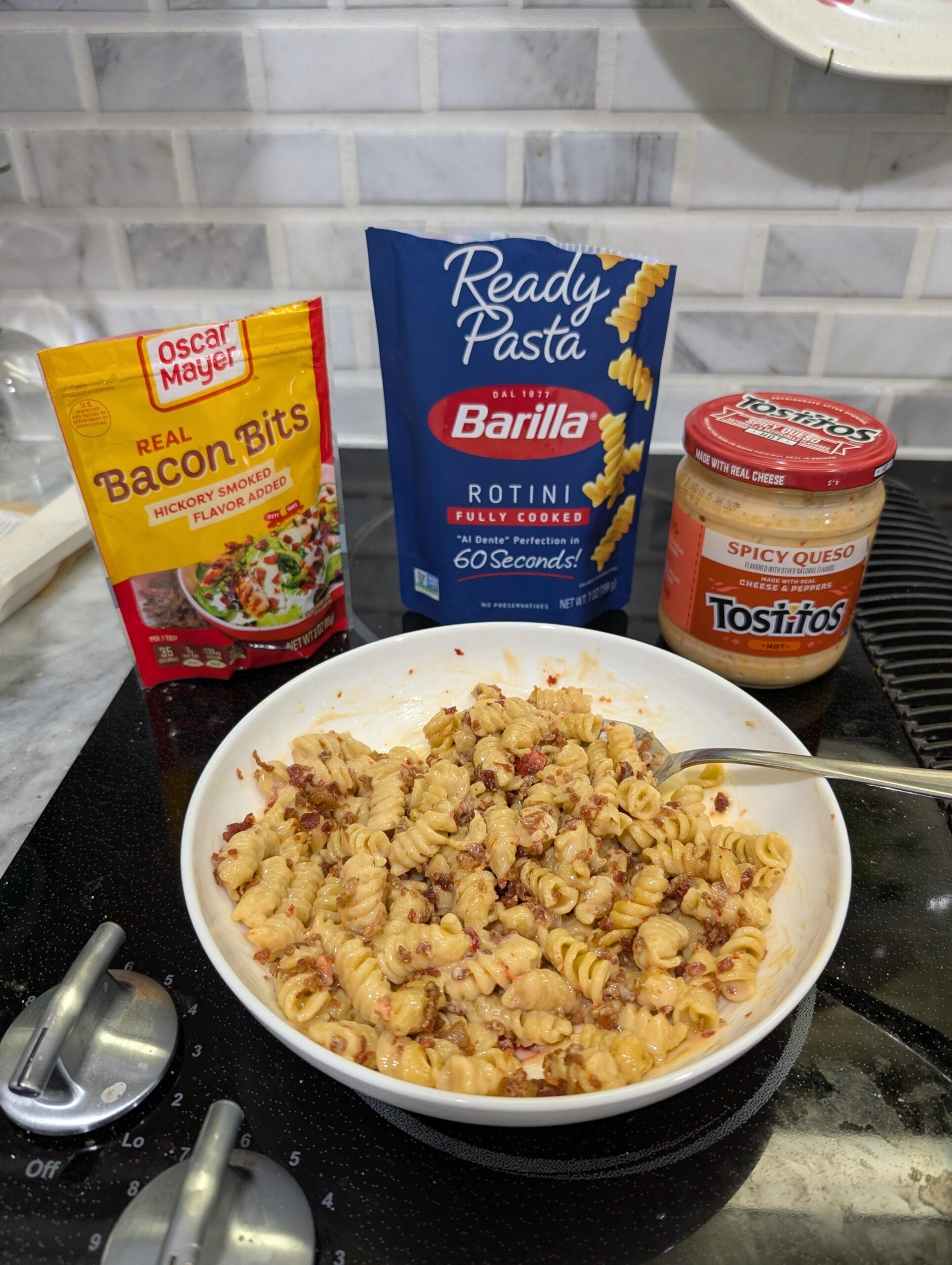 A bowl of rotini pasta mixed with bacon bits and spicy queso sits on a stovetop. Packages of Oscar Mayer Bacon Bits, Barilla Ready Pasta Rotini, and a jar of Tostitos Spicy Queso are in the background.