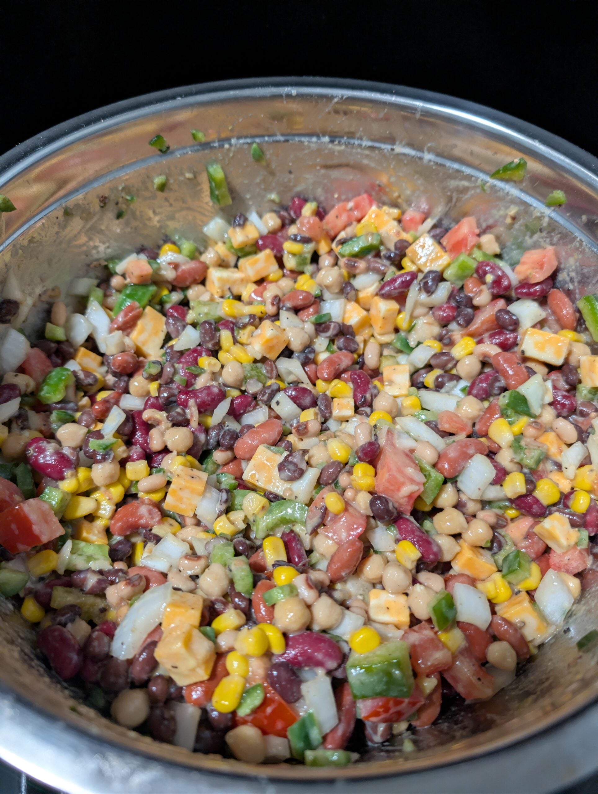 A stainless steel bowl filled with a colorful bean salad containing black beans, kidney beans, chickpeas, corn, diced tomatoes, onions, green bell pepper, and cubes of yellow cheese.