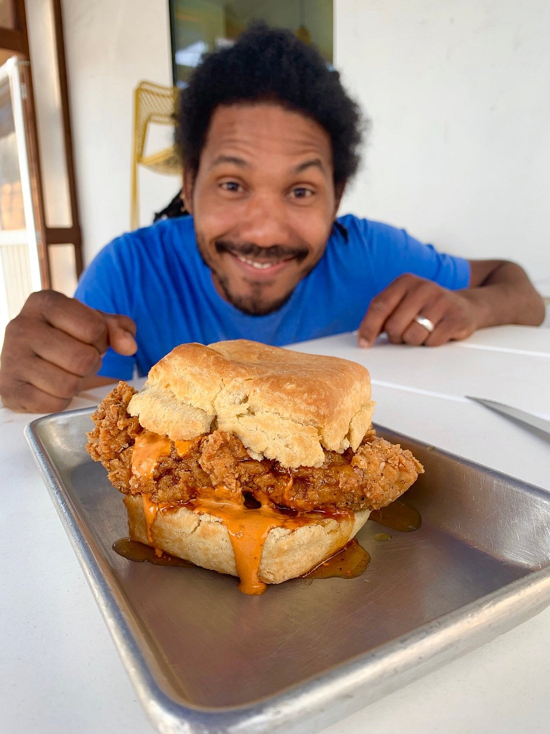 A smiling man in a blue shirt sits at a table, eagerly looking at a biscuit sandwich filled with fried chicken and orange sauce, served on a metal tray.