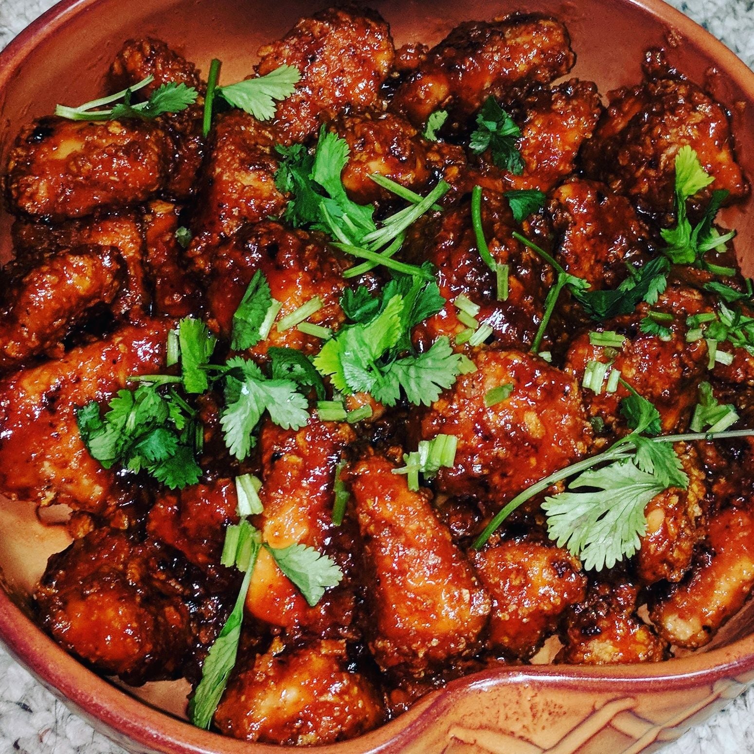 A bowl filled with spicy, saucy chicken wings garnished with fresh cilantro leaves, set against a neutral background.