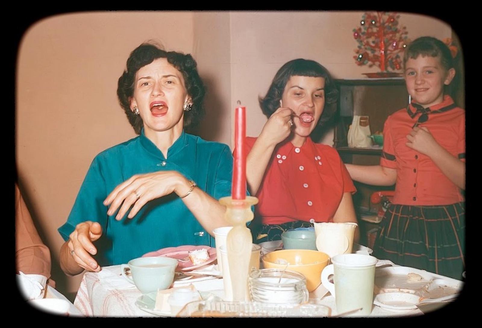 Three people sit at a table with food and dishes, evoking the warmth of nostalgic dinners. Two women are making funny faces while the girl standing beside them smiles. A red candle glows on the table, and a small decorated Christmas tree adds festive charm in the background.