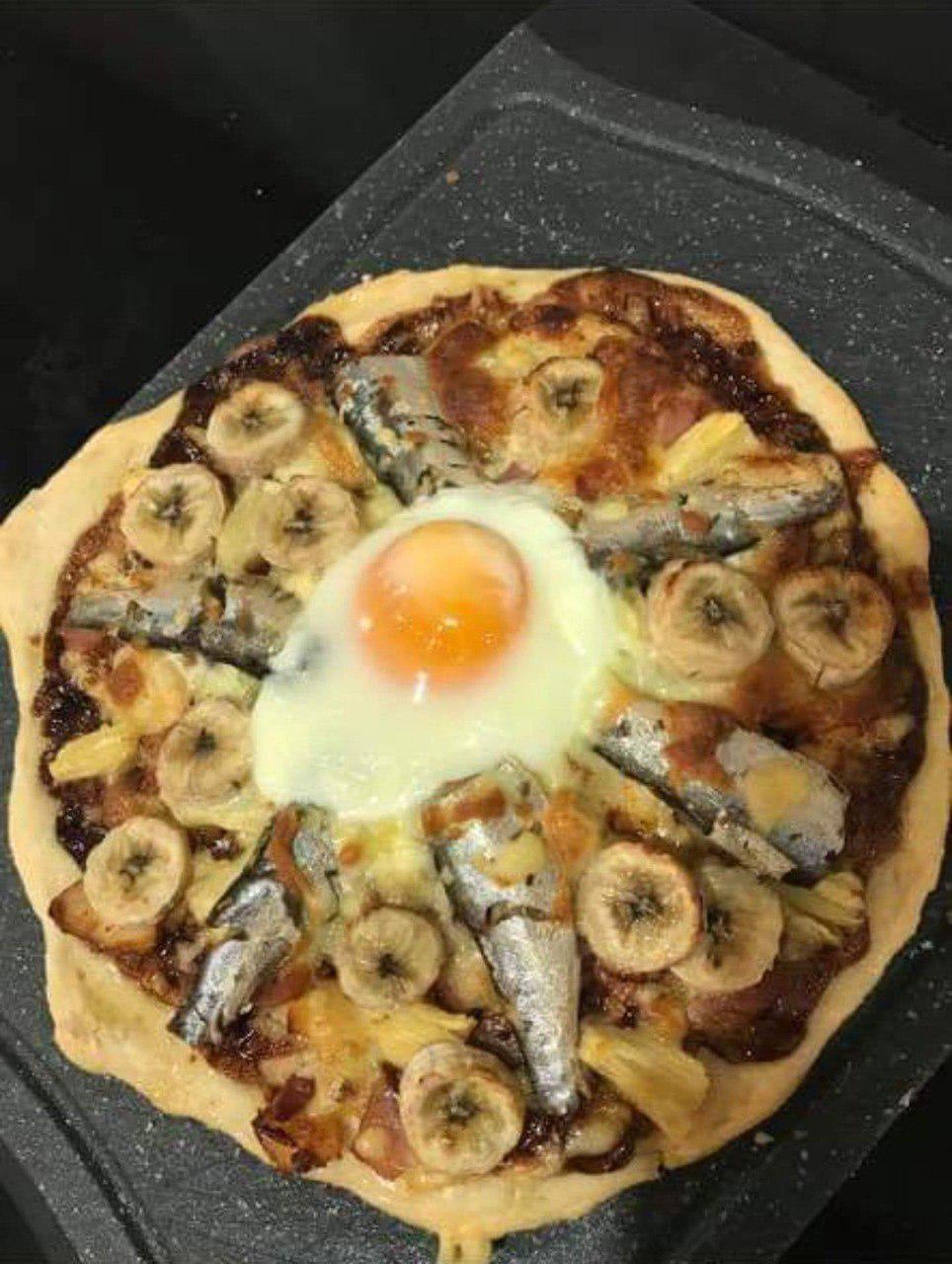 A hilarious cooking disaster comes to life with this pizza, topped with banana slices, sardines, pineapple chunks, and a sunny-side-up egg in the center, all resting on a dark speckled cutting board.