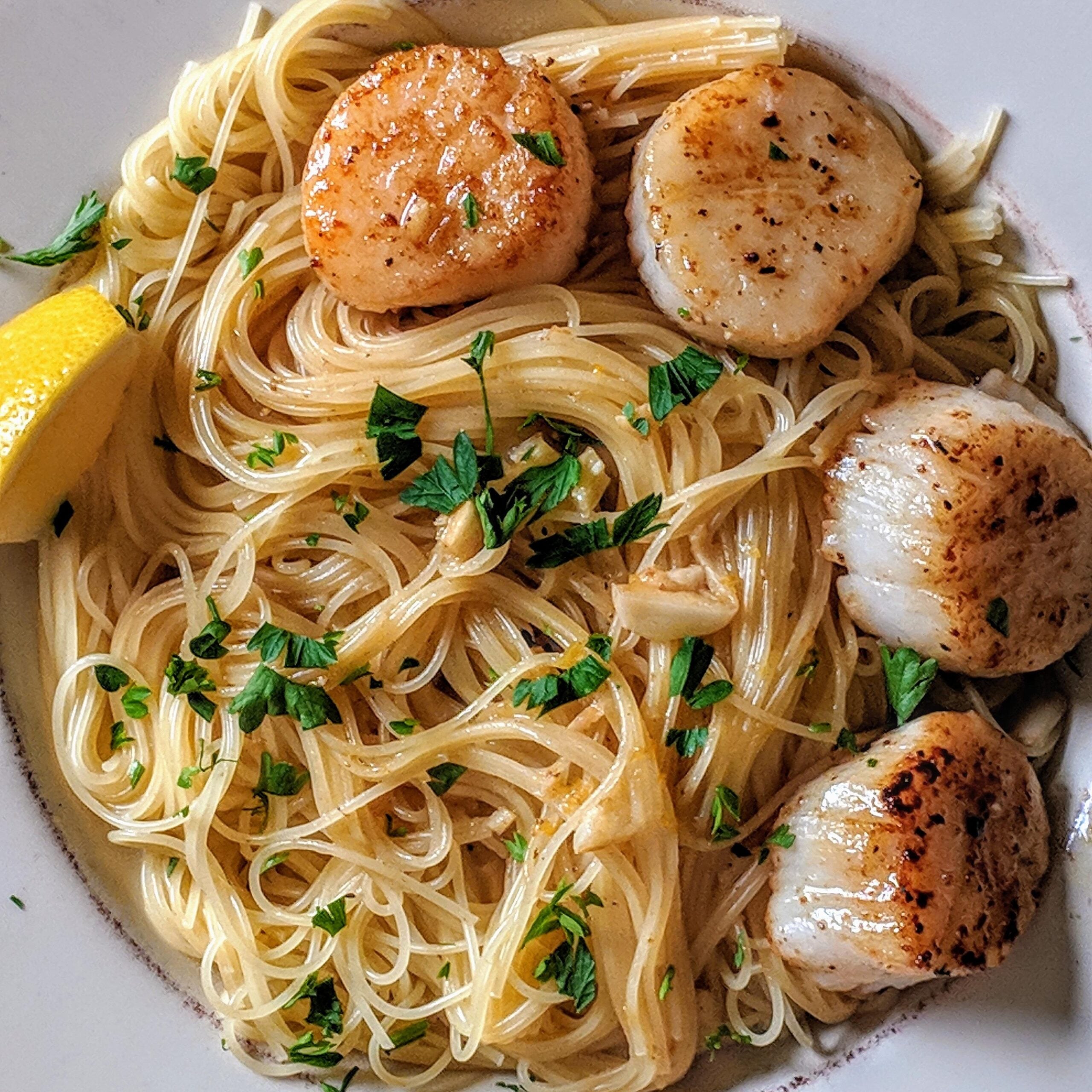 Plate of angel hair pasta topped with four seared scallops, chopped parsley, and a lemon wedge on the side.