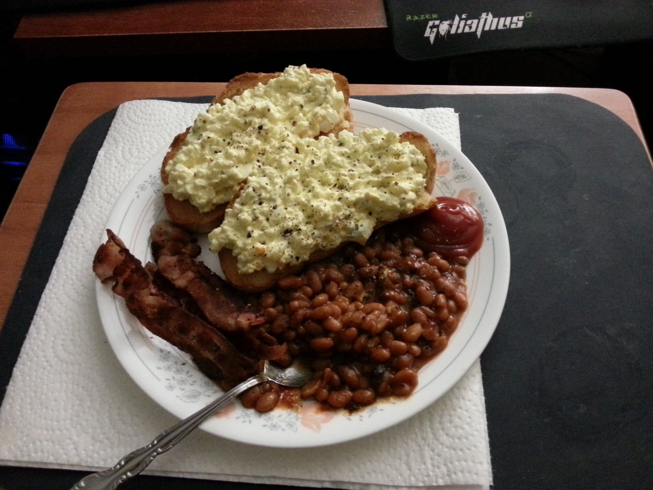 A plate with egg salad on toast, two strips of bacon, baked beans, and a dollop of ketchup. The plate is on a paper towel and a desk, next to a mousepad.