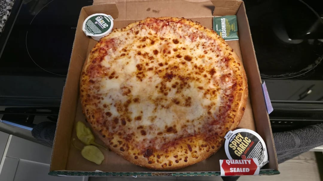 A whole cheese pizza in a cardboard box with two garlic sauce containers, a seasoning packet, and pepperoncini peppers in the corner.