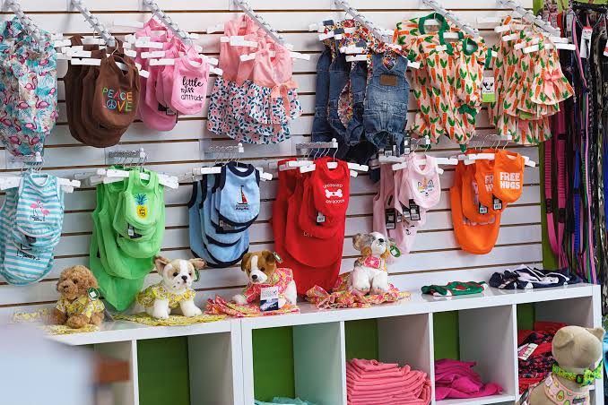 Pet products. Colorful dog clothes hang on a wall display above shelves with plush dog toys and folded garments in a pet store. Leashes are visible on the right side.