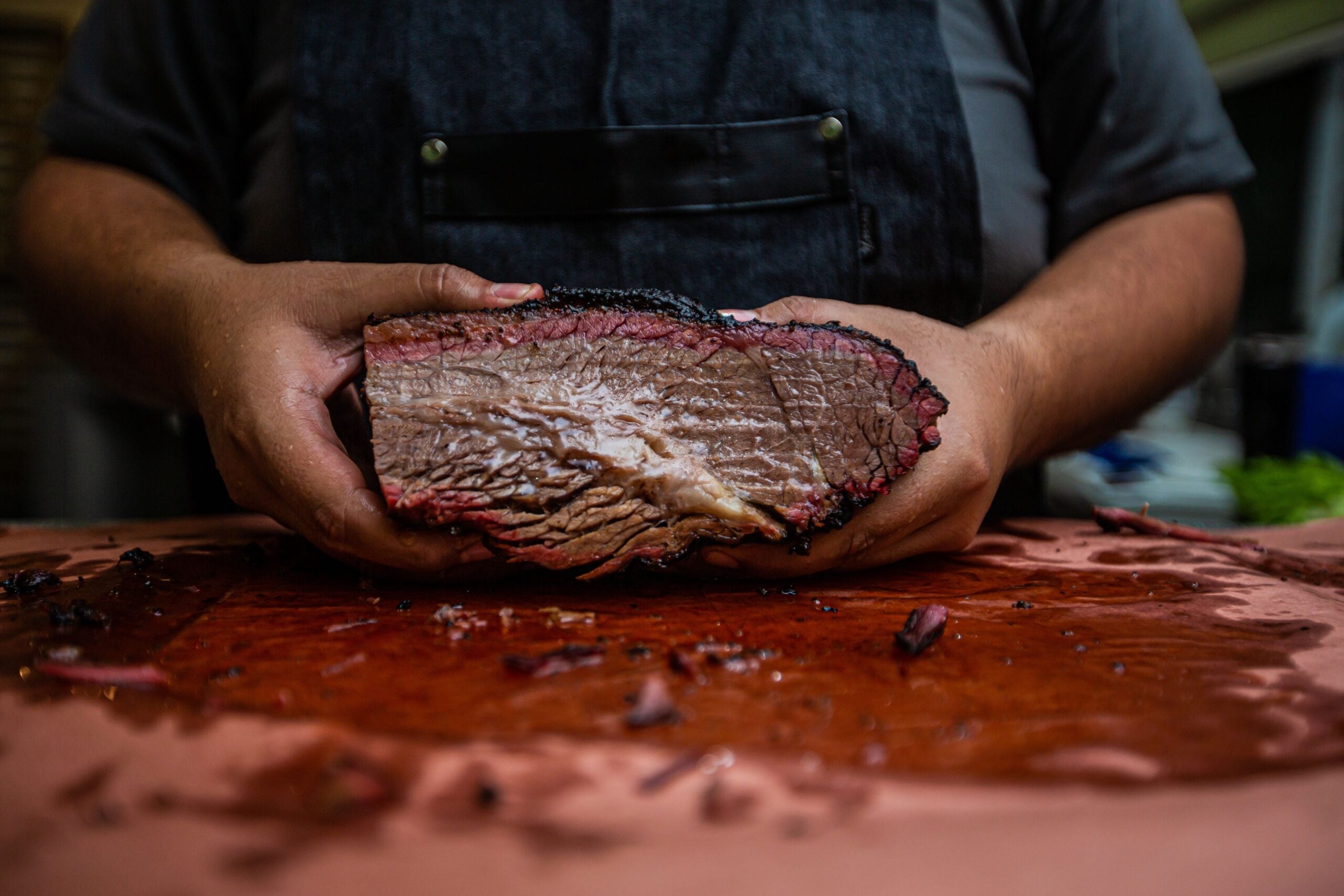 A person in a dark apron holds a large, juicy slice of smoked brisket on a cutting board, showcasing its tender, pink interior and blackened crust.