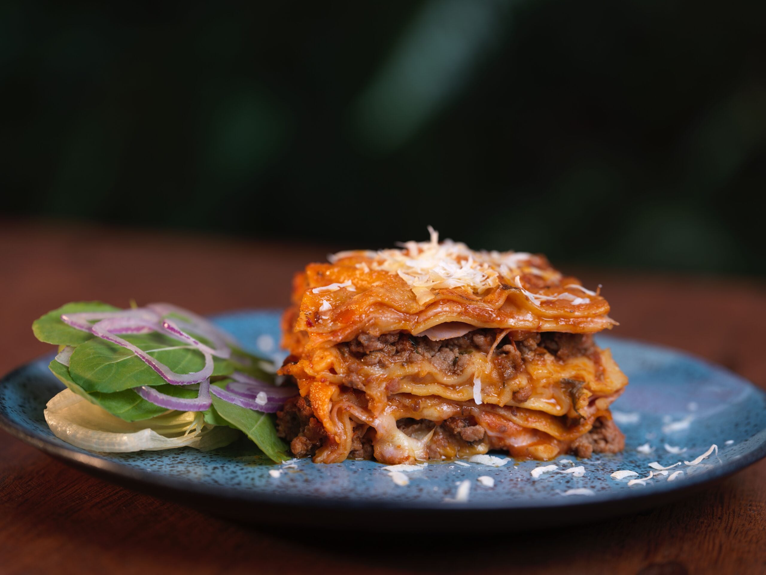 A slice of lasagna with layers of pasta, meat, and cheese sits on a blue plate, garnished with grated cheese. A small side salad with spinach, red onion, and cucumber is next to it.