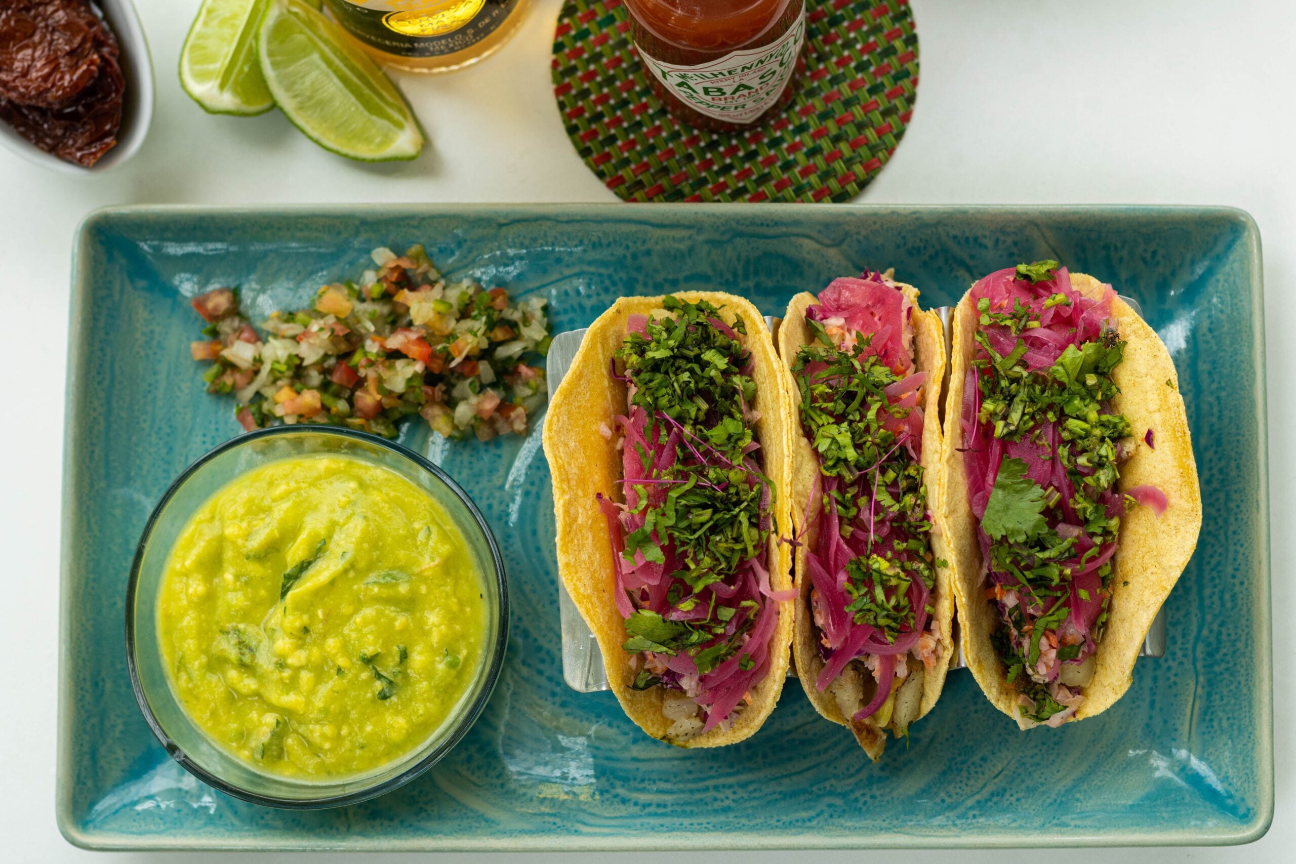 Three tacos filled with onions and herbs on a blue plate, served with a side of green sauce and pico de gallo. Lime wedges, hot sauce, and a drink are partially visible in the background.