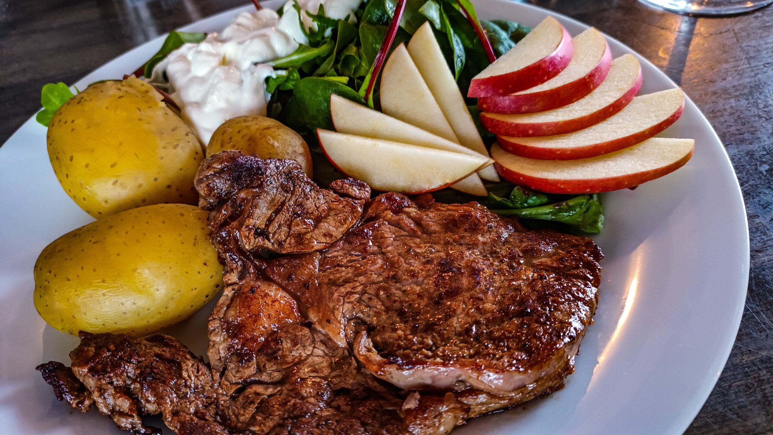 A plate with grilled steak, three boiled potatoes, fresh green salad with dressing, and thinly sliced red apples.