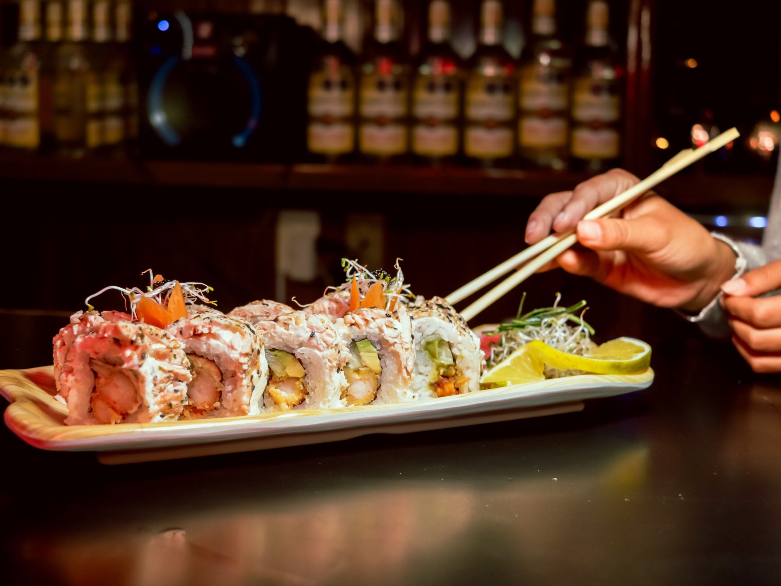 A hand uses chopsticks to pick up a sushi roll from a rectangular plate, garnished with greens, lemon slices, and sauce. Bottles are blurred in the background on a dimly lit bar.