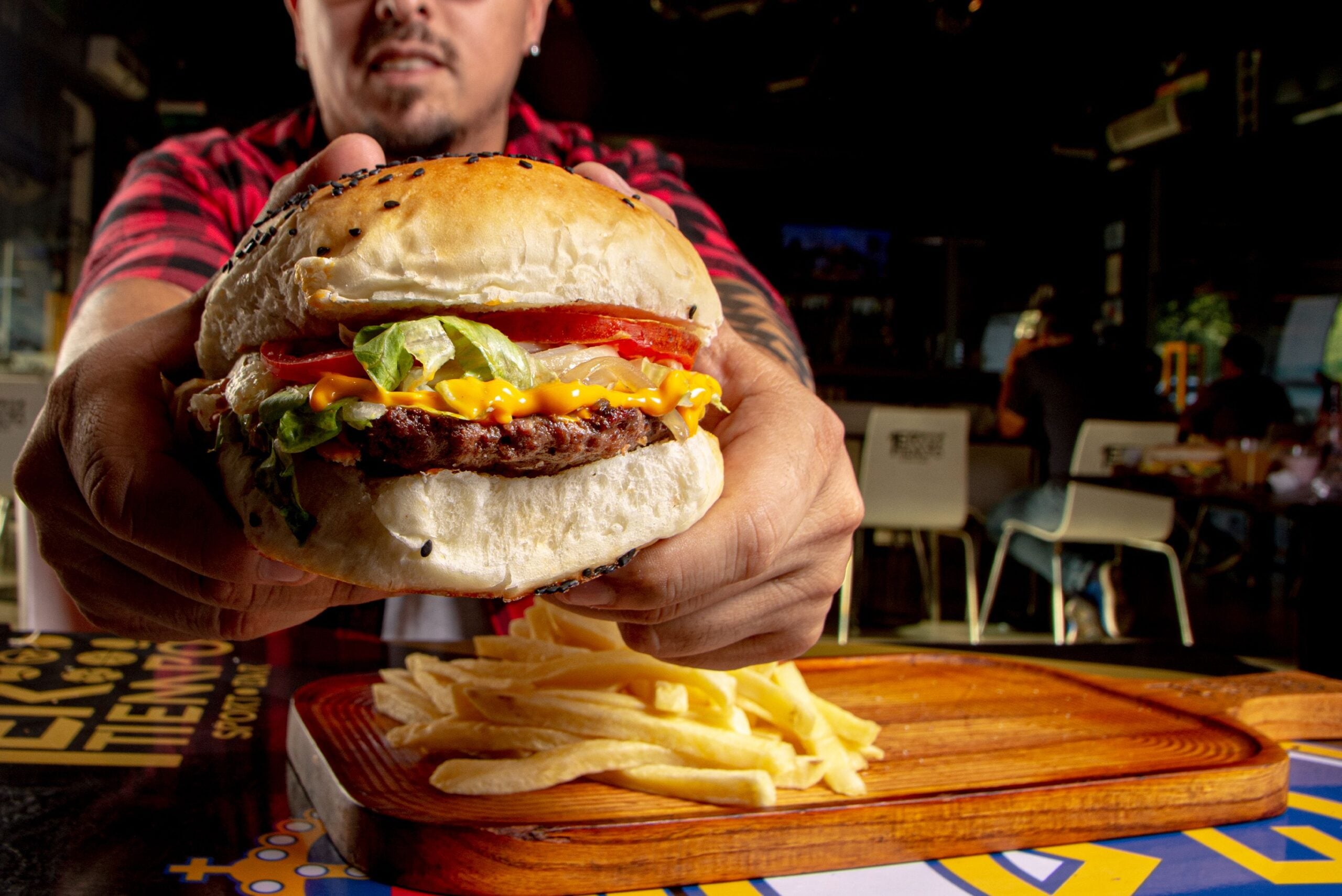 A person in a red plaid shirt holds a large burger with lettuce, tomato, and cheese toward the camera. A serving of French fries sits on a wooden tray on the table in front.