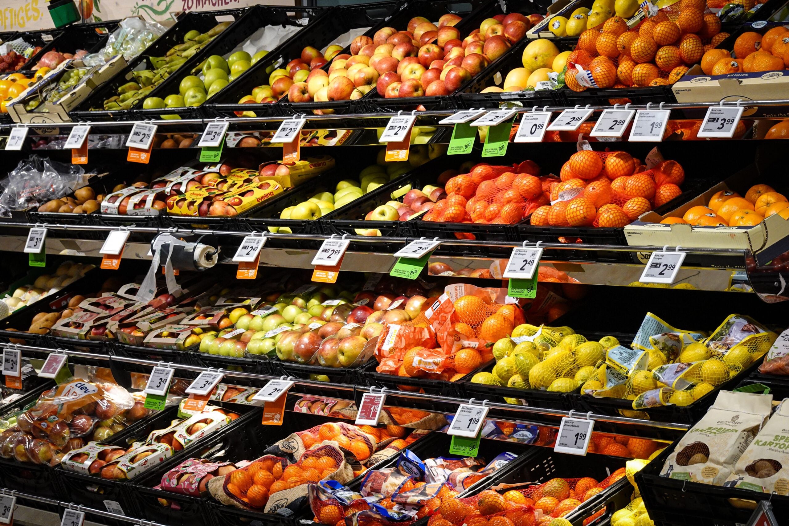 A grocery store produce section with neatly stacked apples, oranges, lemons, pears, and other fruits, each labeled with price tags and separated in black trays on slanted shelves.
